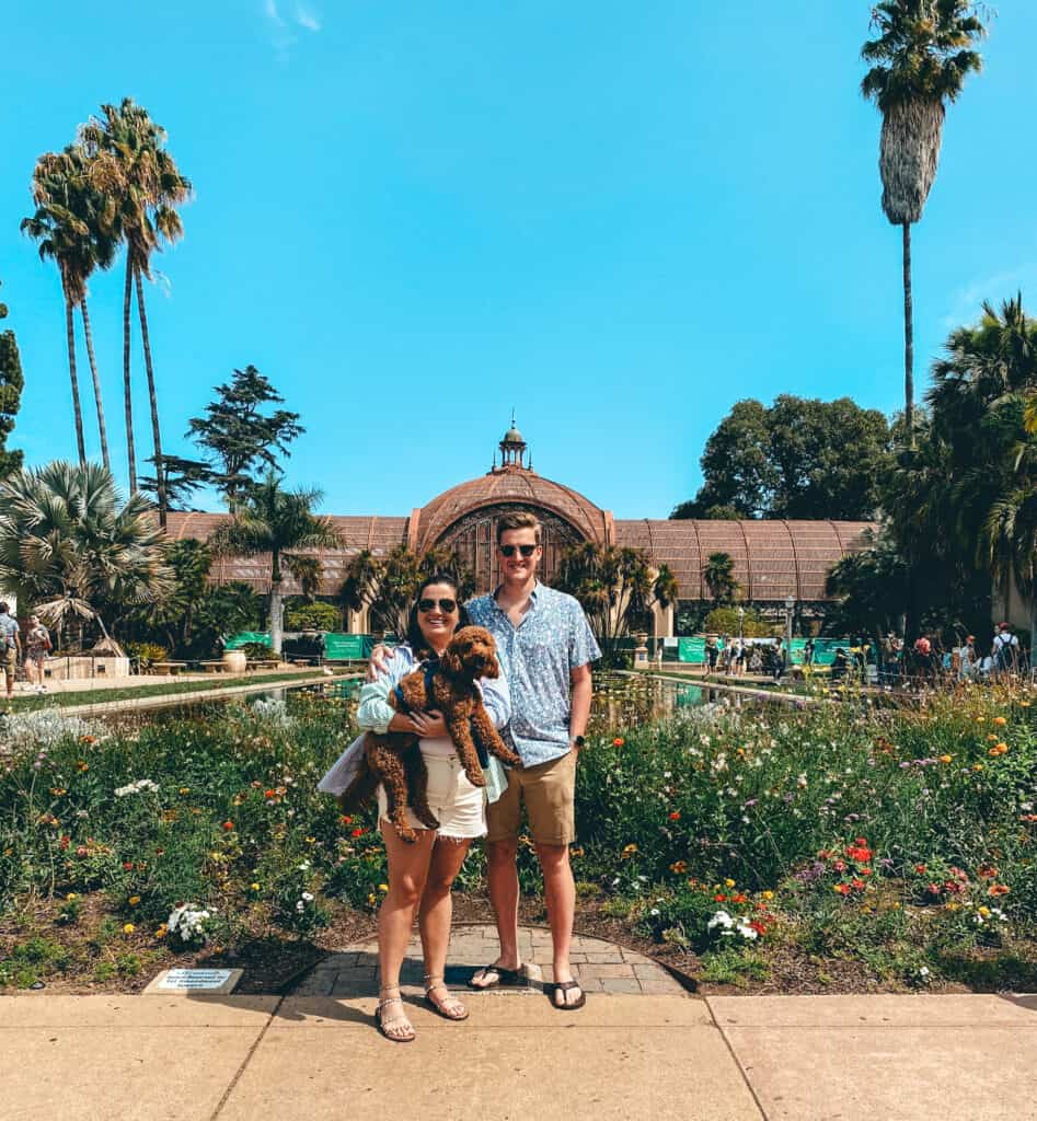 A smiling couple stands in front of the botanical building in Balboa Park, San Diego, holding their brown poodle. Lush gardens and towering palm trees under a clear blue sky create a scenic backdrop for the happy moment.