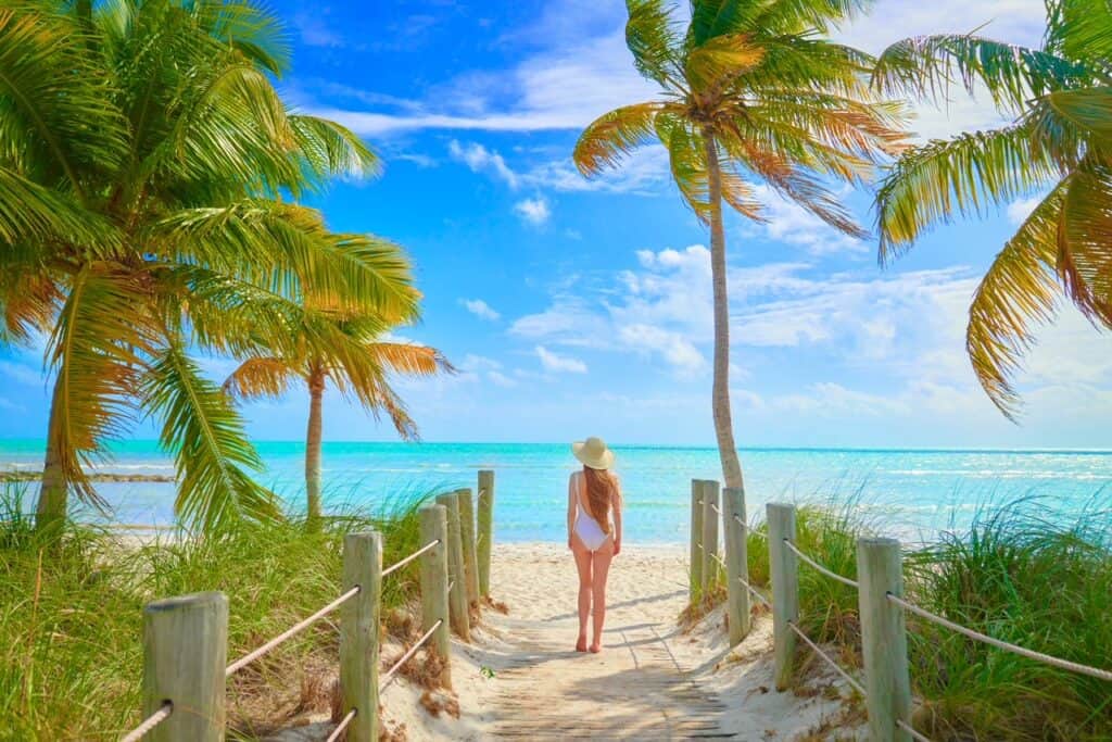 A woman in a sunhat and white dress gazes out to the azure waters of the Florida Keys, framed by lush palm trees and a sandy beach path.