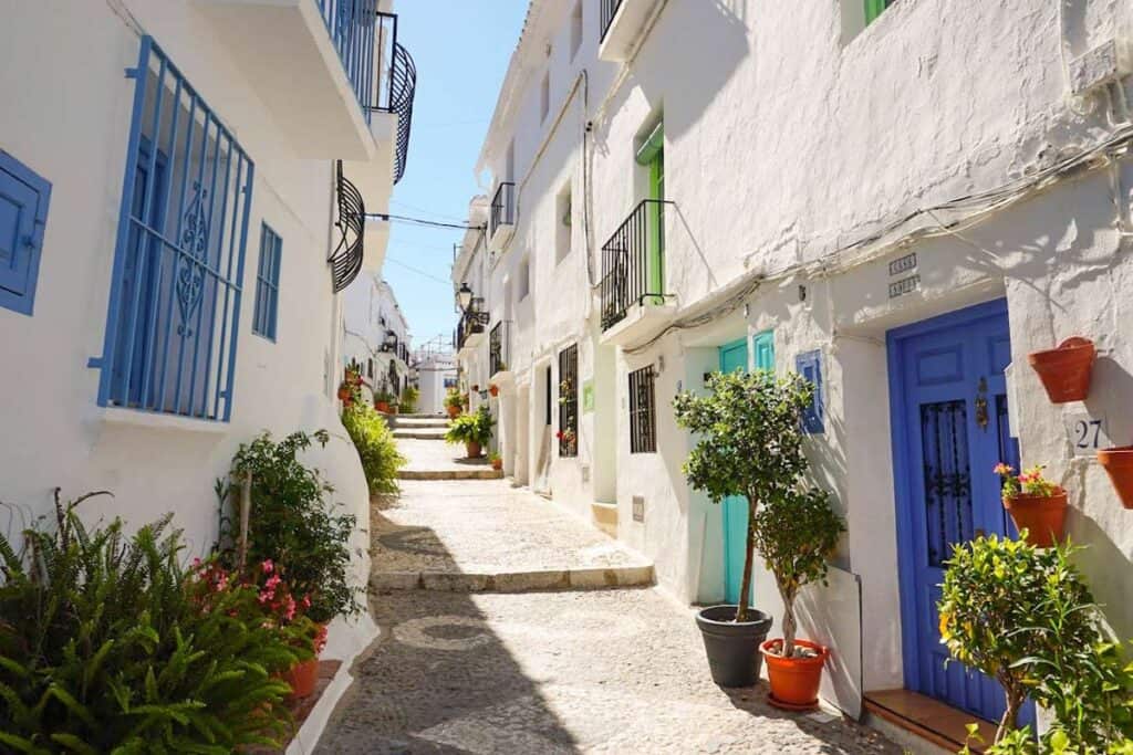 The image shows a picturesque street in Frigiliana, Spain, lined with traditional white-washed houses adorned with colorful doors and window frames. Potted plants and blooming flowers add a vibrant touch to the charming cobblestone pathway, invoking a sense of Mediterranean warmth and hospitality.