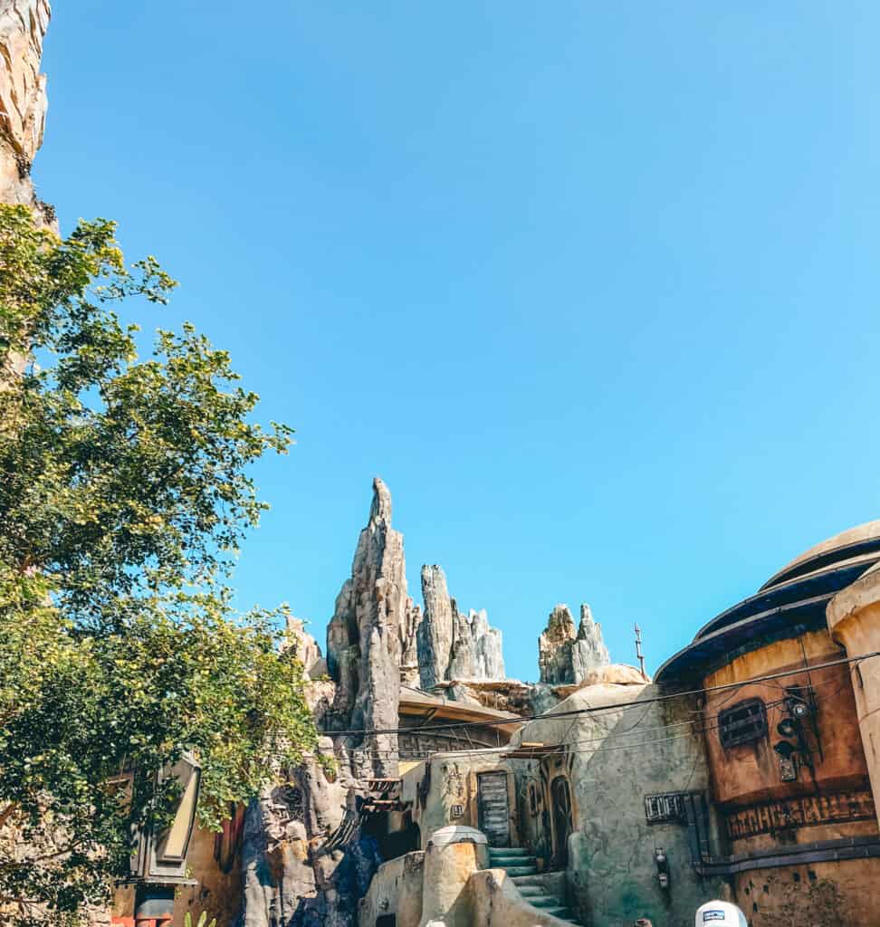 A vivid photo capturing the rugged terrain of Star Wars: Galaxy's Edge at Disneyland. Jagged rock formations tower above intricately detailed, weathered buildings reminiscent of a bustling spaceport, all set against a bright blue sky.