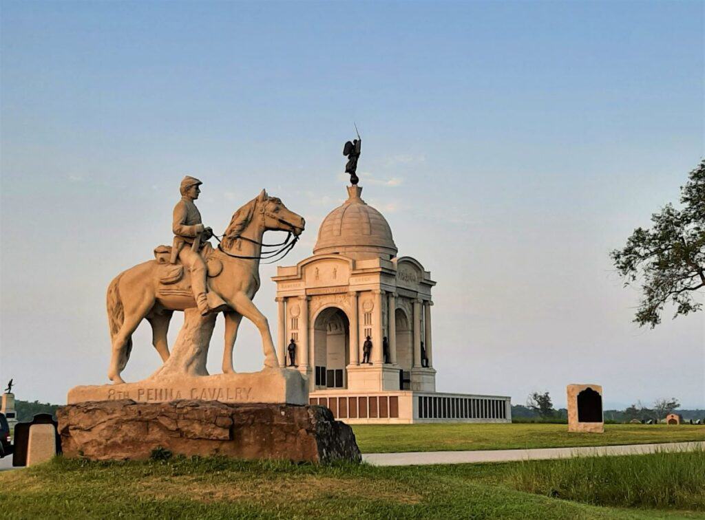 Early evening view of the Pennsylvania State Memorial at Gettysburg National Military Park with a foreground statue of a soldier on horseback. The monument's classical dome and columns are illuminated by the warm hues of a setting sun.