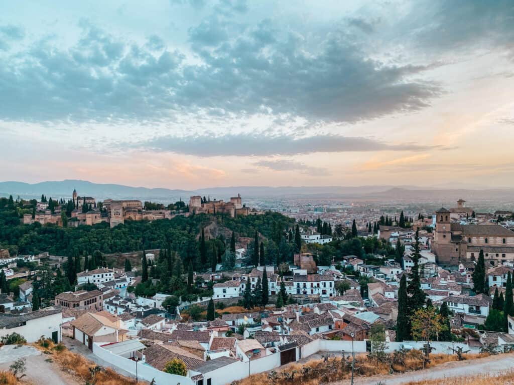 The image features a panoramic view of Granada, Spain, during twilight. The Alhambra, a famous palace and fortress, dominates the landscape with its ancient walls and palatial structures surrounded by lush greenery, set against a backdrop of a soft sky with hues of pink and blue, overlooking the city with traditional white houses below.