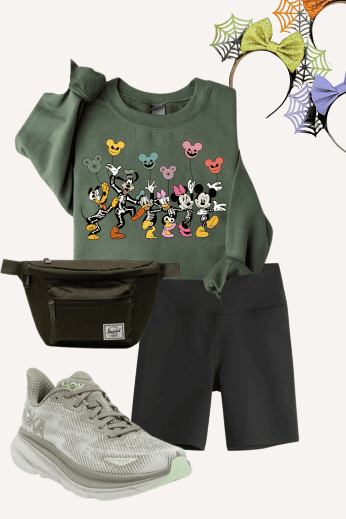 A playful and colorful Halloween outfit arrangement. Centered is an olive green sweatshirt adorned with classic cartoon characters dressed as skeletons. A dark pair of athletic shorts and a green waist bag are positioned below. The outfit is finished with a pair of light green and white running shoes and a Halloween-themed mouse ears headband with cobweb design and multicolored bows.