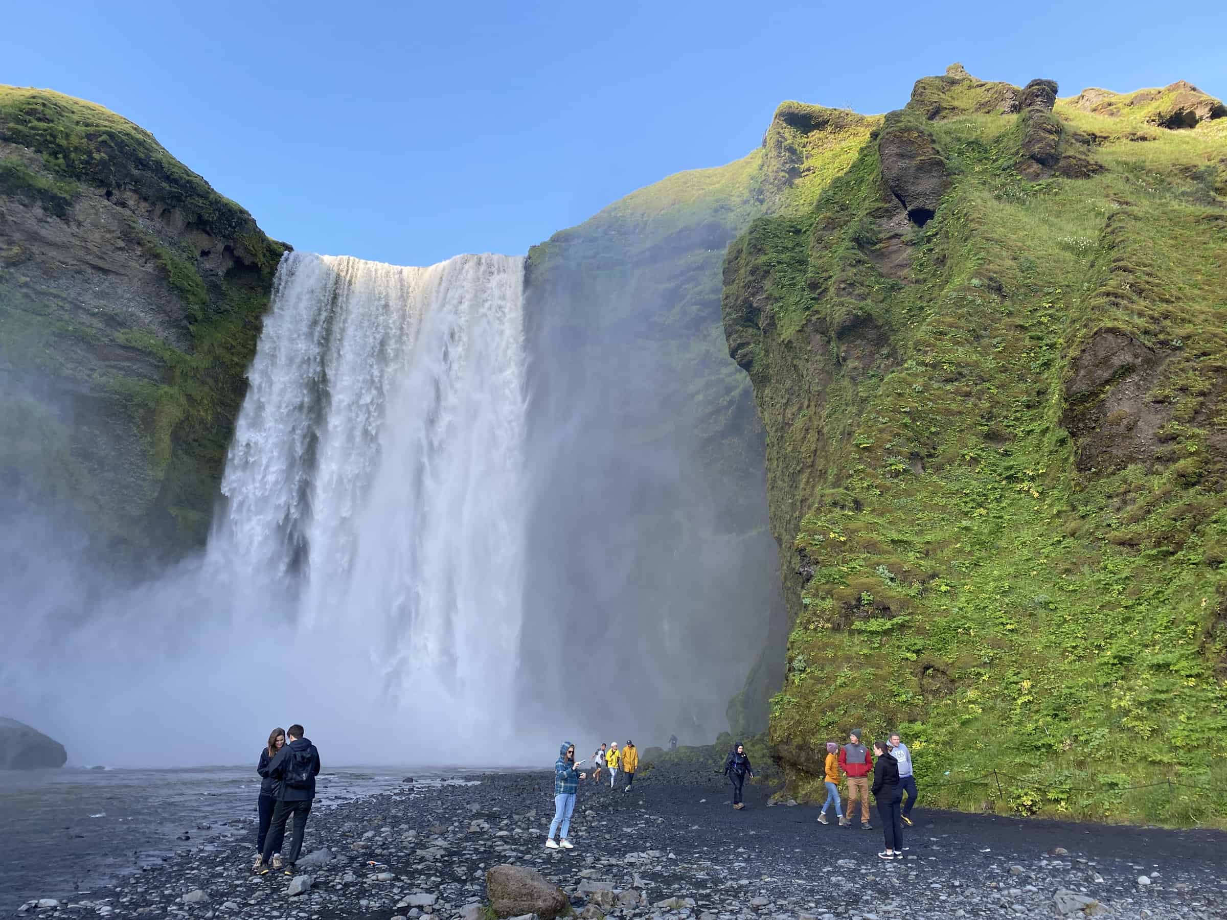 Tourists enjoying the magnificent Skógafoss waterfall in Iceland, with its powerful cascade plummeting down a green moss-covered cliff into a misty pool below, against a backdrop of a clear blue sky
