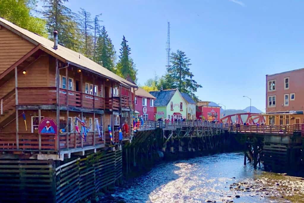 Quaint creek-side boardwalk with colorful buildings in Ketchikan, Alaska. The wooden structures and vibrant facades reflect the town's historic charm against a backdrop of evergreen trees and a clear blue sky.