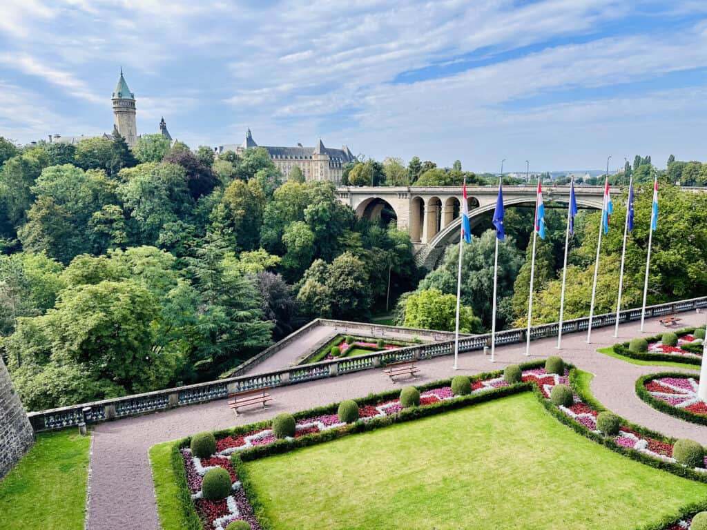 The image presents a verdant view of Luxembourg City, with a focus on the meticulously maintained gardens featuring geometric flower beds. In the background, the iconic Adolphe Bridge arches over a lush gorge, while European and Luxembourgish flags line a promenade under a blue sky with fluffy clouds.