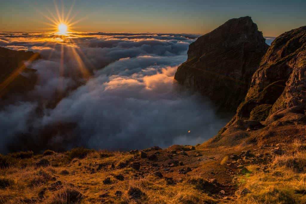 The image depicts a majestic sunrise over the mountains of Madeira, Portugal. The sun bursts through the clouds, casting a golden glow over the rugged terrain and the sea of clouds nestled in the valleys, creating a dramatic and ethereal landscape.