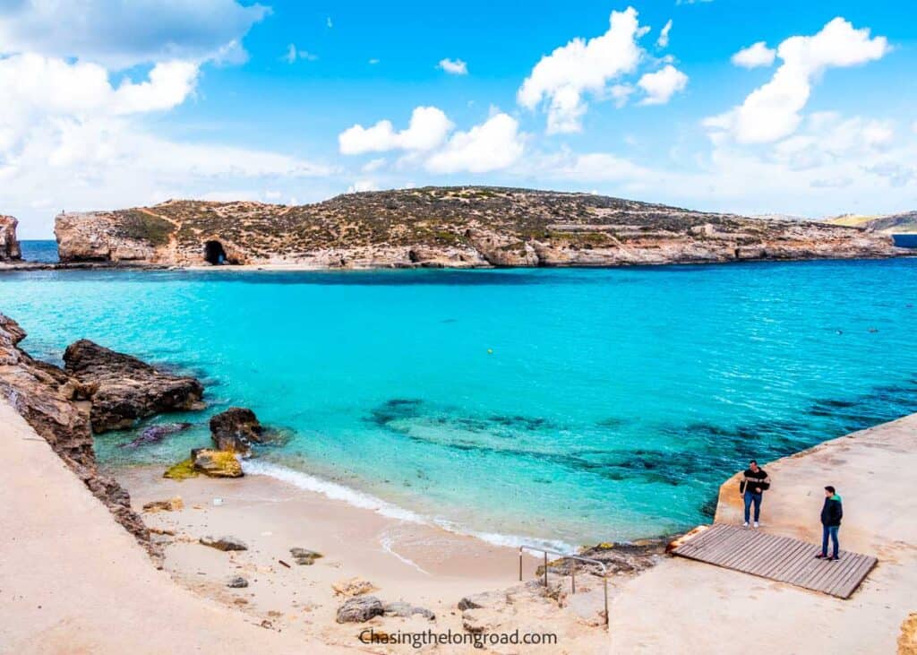 The image captures a striking view of the Blue Lagoon in Malta, with its vivid turquoise waters surrounded by rugged limestone cliffs. Two individuals stand at the water's edge on a concrete jetty, enhancing the scale and natural beauty of the serene coastal setting. The website 'Chasingthelongroad.com' is watermarked at the bottom, suggesting travel or adventure content.