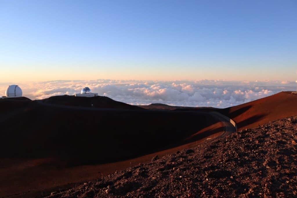 The observatories atop Mauna Kea on the Big Island of Hawaii bask in the warm glow of sunset, with the summit road winding across the barren volcanic landscape above the cloud line