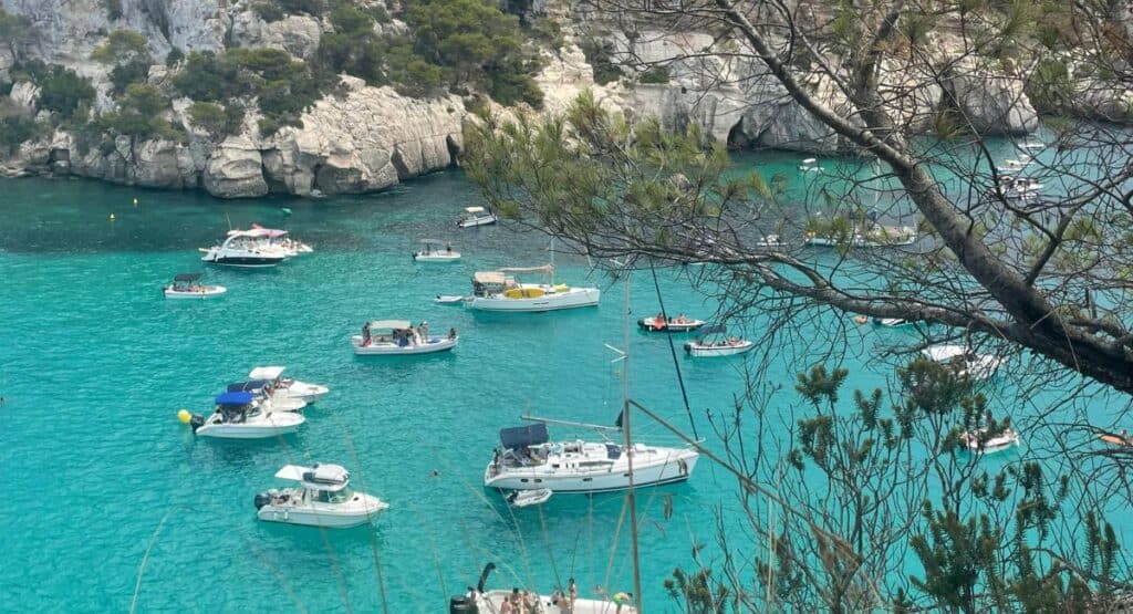 The image captures a scenic view of the turquoise waters of a cove in Menorca, Spain, dotted with various boats and yachts. The lush greenery of pine trees partially frames the top of the view, with rocky cliffs enclosing the serene bay, creating a peaceful Mediterranean seascape.