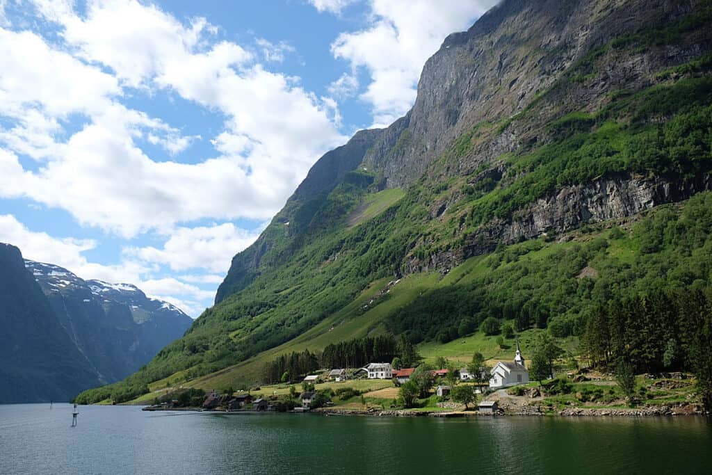 The tranquil waters of Nærøyfjord in Norway, with a small village nestled at the base of steep green cliffs, a traditional white church standing out amidst the natural scenery, under a sky with scattered clouds.