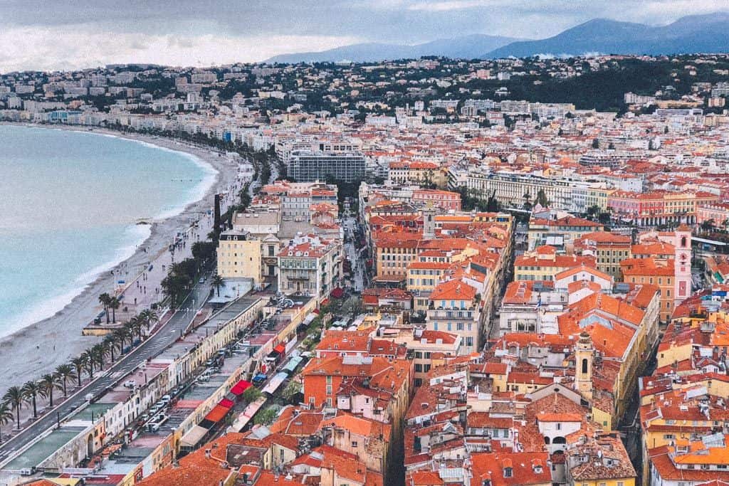 The image provides an aerial view of Nice, France, capturing the dense, colorful buildings of the city juxtaposed with the curved Bay of Angels coastline. The Promenade des Anglais is visible as a sweeping boulevard lined with palm trees, bordering the pebbly shores of the Mediterranean Sea, with overcast skies above the urban landscape.