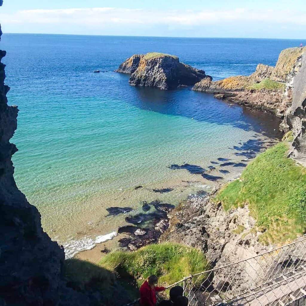 A secluded cove off the coast of Northern Ireland, with clear turquoise waters enclosed by rugged cliffs and rocky outcrops, accessible by a steep staircase with visitors enjoying the hidden gem under a clear blue sky.
