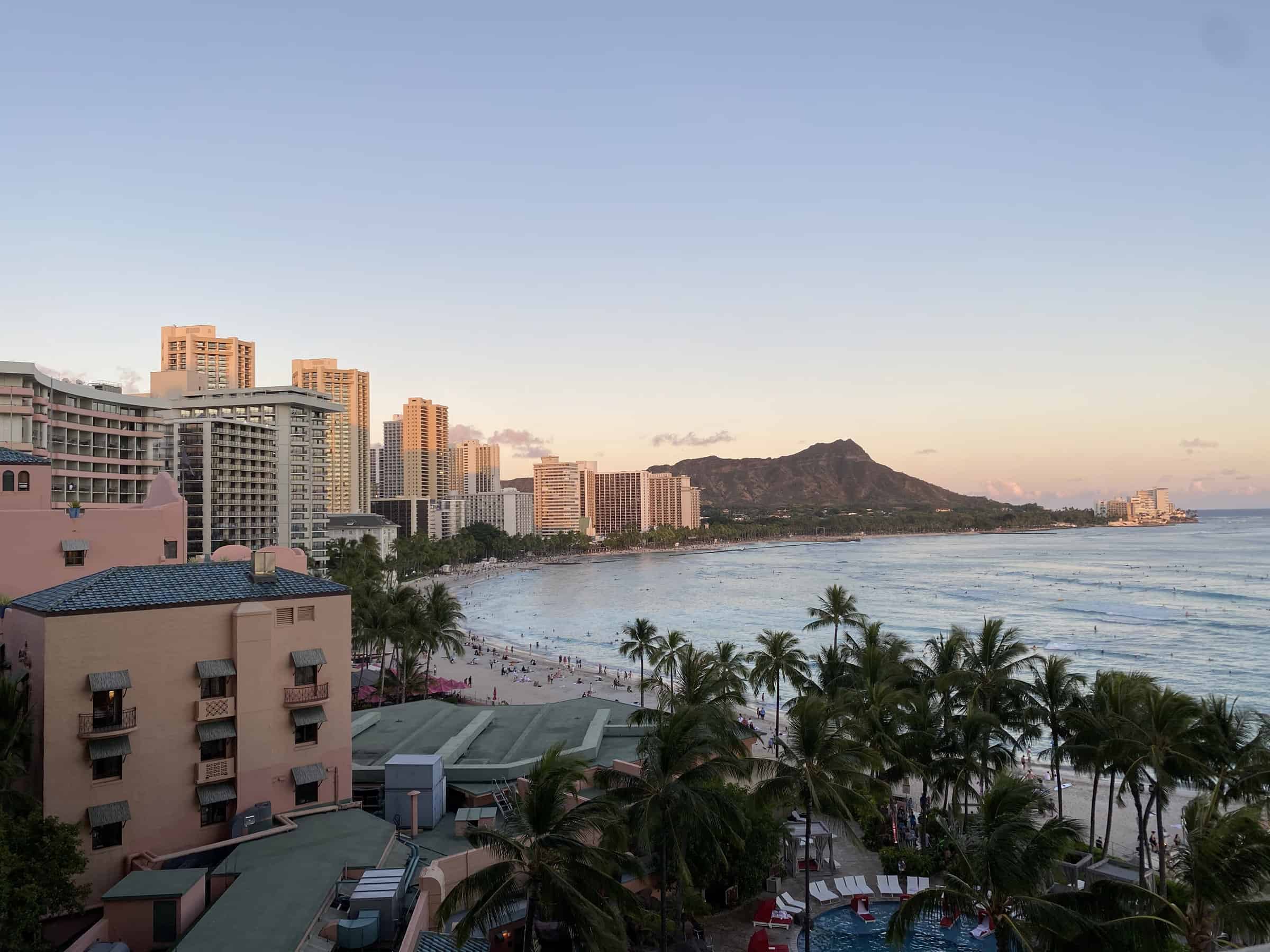 Scenic view of Waikiki Beach in Oahu, Hawaii, at dusk with Diamond Head Crater in the distance. The coastline is lined with palm trees, sandy beaches, and a series of high-rise hotels against a soft pastel sky.