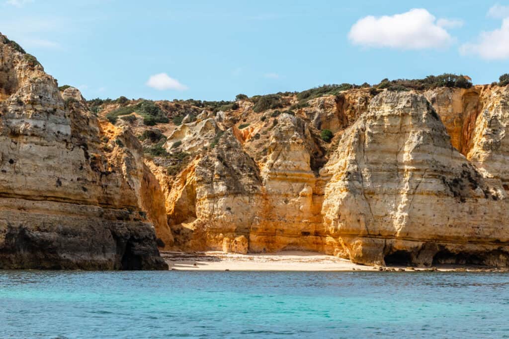 The stunning cliffs of Lagos in the Algarve region of Portugal, with their natural golden rock formations rising from clear turquoise waters, and a secluded sandy beach nestled between the cliffs under a bright blue sky.