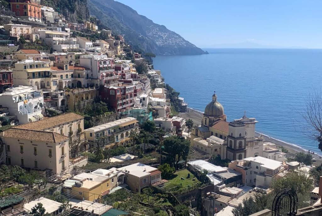 The cliffside village of Positano on Italy's Amalfi Coast, with its colorful houses and the majestic Santa Maria Assunta church featuring a tiled dome, overlooking the serene blue Tyrrhenian Sea on a sunny day.