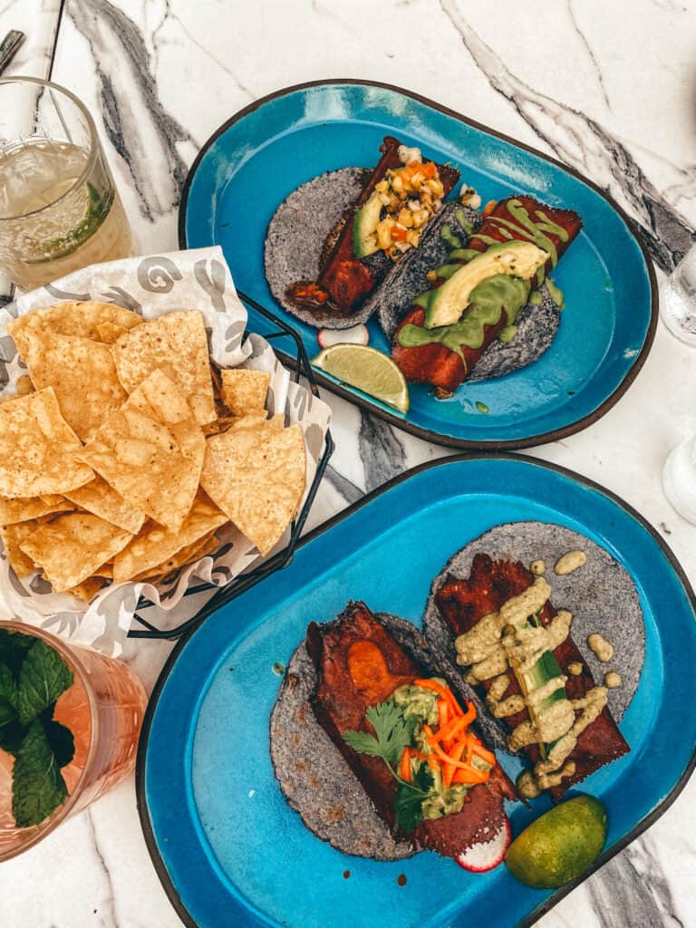 A top-down view of a vibrant Mexican meal at Puesto, featuring blue corn tortillas with an assortment of tacos, fresh avocado slices, and a tangy corn salsa. Accompanied by a basket of crispy tortilla chips and refreshing drinks on a marble table.