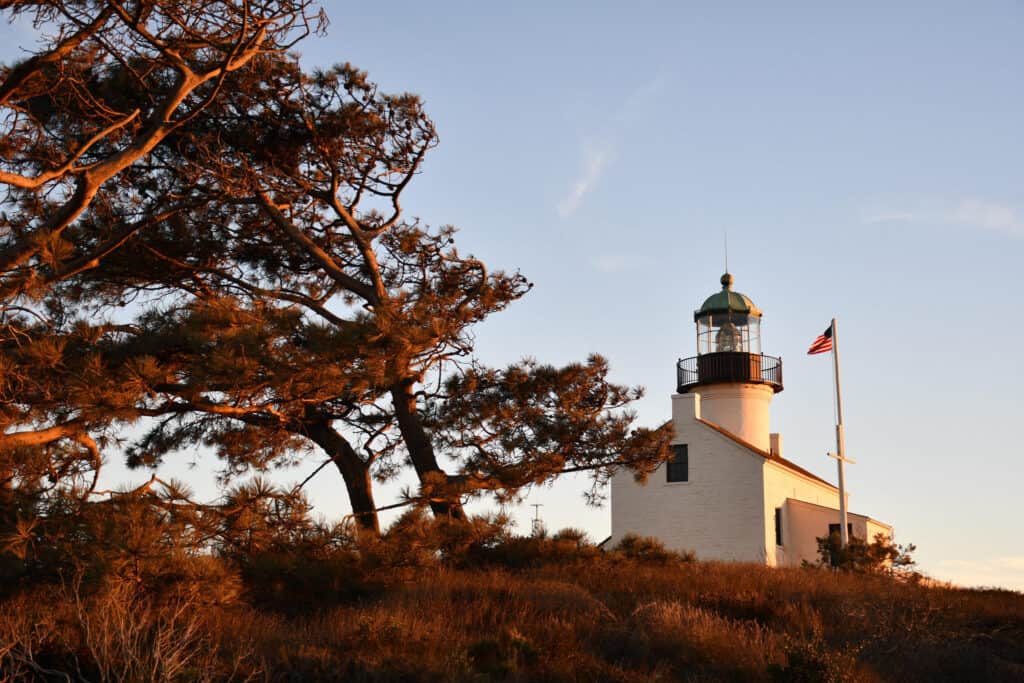 The historic Point Loma Lighthouse in San Diego, California, captured at golden hour with a backdrop of a twisted pine tree and the American flag waving in the breeze.