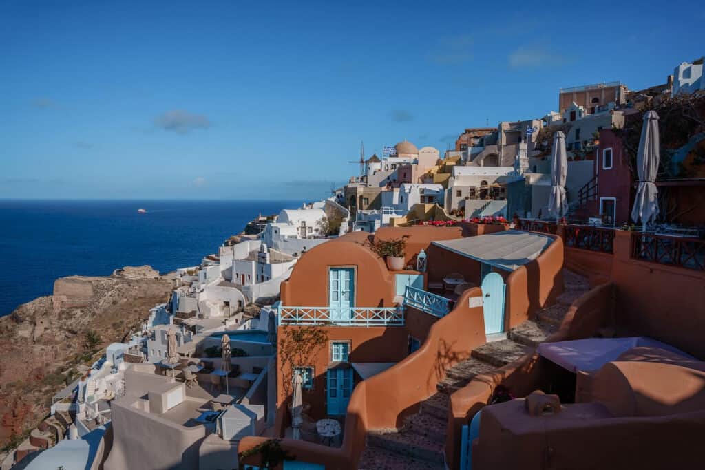 The image showcases the iconic cliffside architecture of Santorini, Greece, with its cascading white buildings and blue accents, overlooking the serene Aegean Sea. The clear blue sky above and the calm sea below frame the unique Cycladic architecture beautifully, capturing the essence of this popular Mediterranean destination.