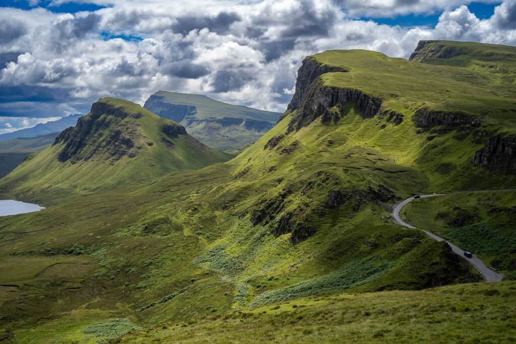 Dramatic landscape of the Quiraing on the Isle of Skye, Scotland, with winding roads cutting through lush green hills and rugged cliffs, under a dynamic sky with patches of sunlight.