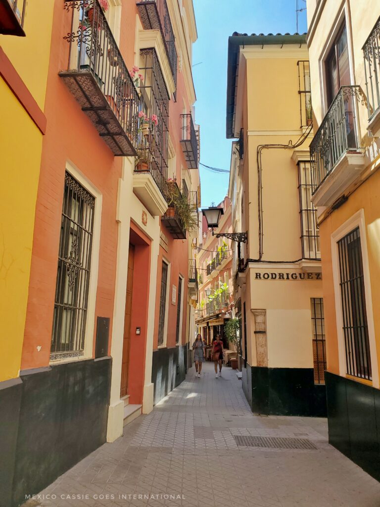 The image features a narrow, sunlit street in Seville, Spain, lined with colorful buildings and hanging plants on the balconies. Two people are seen walking down the street, contributing to the lively, urban atmosphere of the city. 
