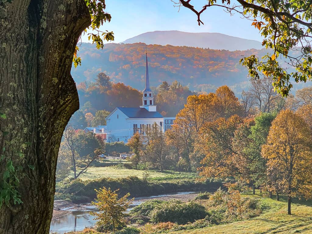 A tranquil morning view of the Stowe Community Church in Vermont, its white steeple standing tall against a backdrop of colorful autumn trees and a gentle river