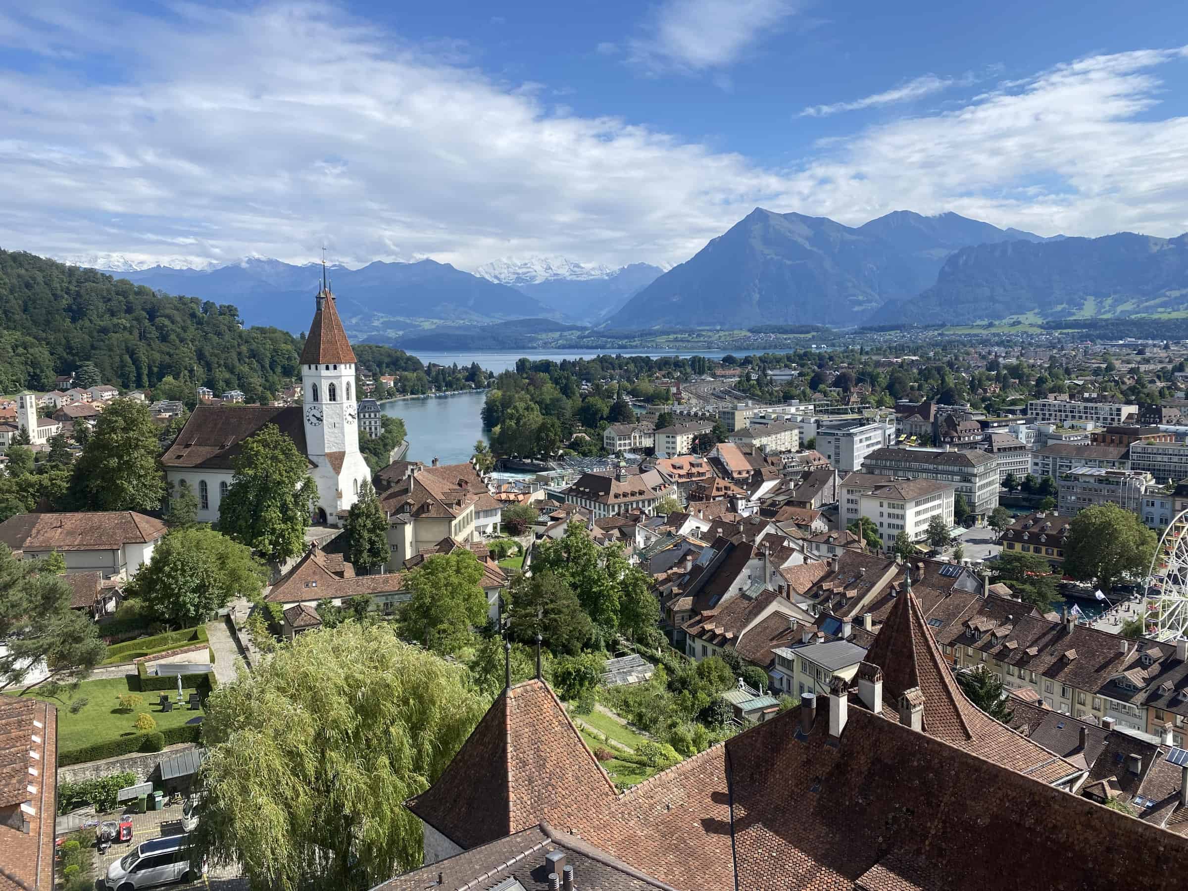 Panoramic view of Thun, Switzerland, with the historic Scherzligen Church in the foreground, overlooking the serene blue waters of Lake Thun, framed by the Swiss Alps in the distance on a partly cloudy day