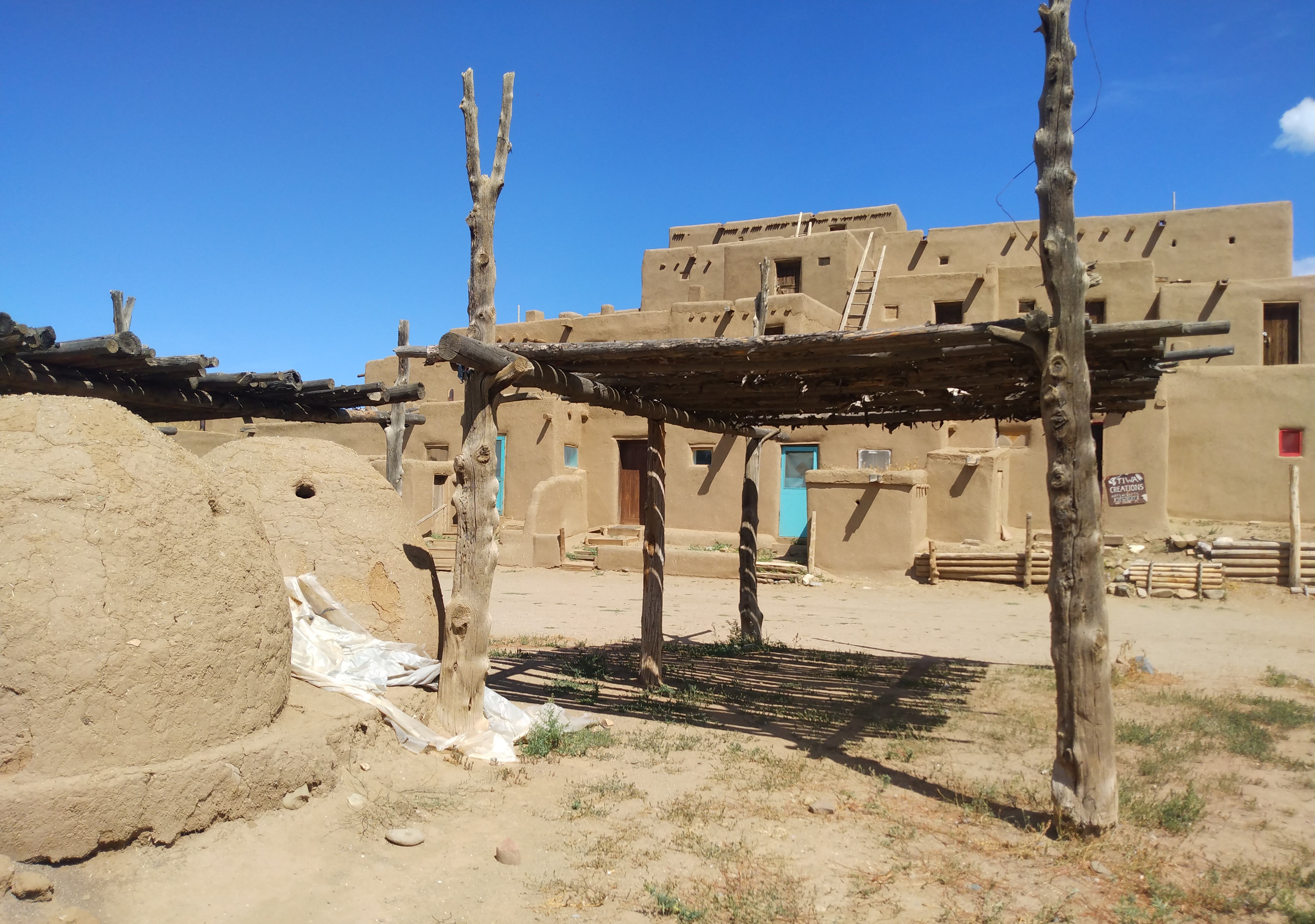 Traditional adobe buildings at the Taos Pueblo in New Mexico under a clear blue sky. The ancient pueblo structures feature wooden ladders, earthen walls, and are a testament to the enduring Native American architecture