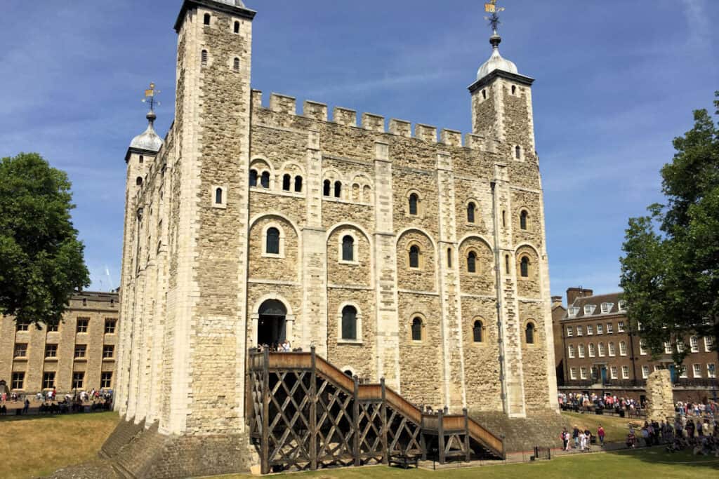 The Tower of London on a sunny day, with visitors gathered around the historic fortress's White Tower, which showcases its medieval architecture, including the stone walls and the wooden drawbridge entrance.