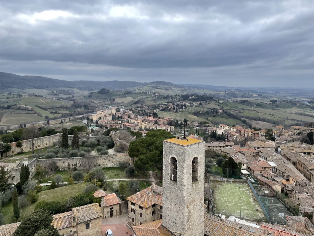 The image captures an overcast day overlooking a picturesque Tuscan landscape in Italy. In the foreground, there's a stone tower rising above terracotta-roofed buildings, while the background reveals rolling hills dotted with homes, cypress trees, and farmlands, all under a dramatic grey sky.