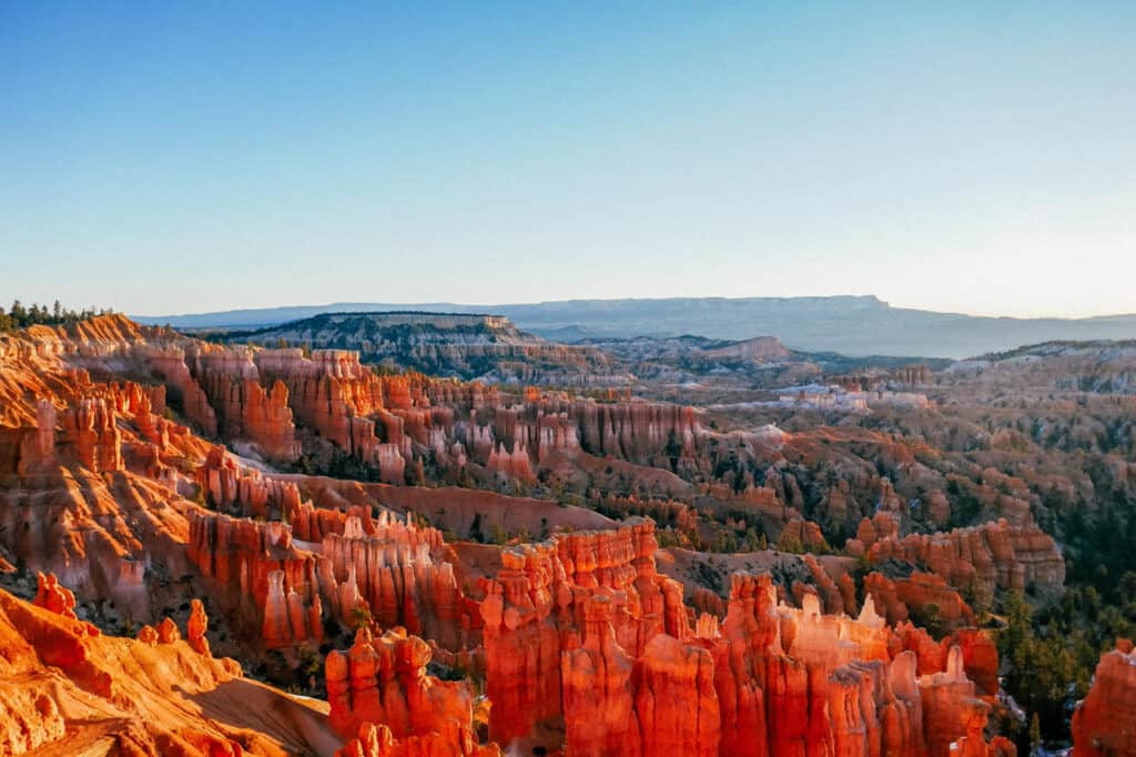 The warm glow of sunrise illuminates the red rock hoodoos of Bryce Canyon National Park in Utah, highlighting the intricate formations and vast, rugged landscape.