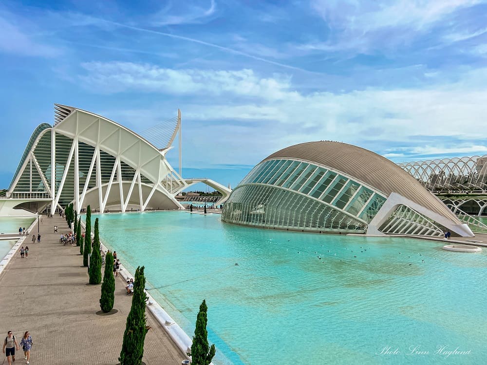 The image presents a panoramic view of the City of Arts and Sciences in Valencia, Spain. It features futuristic architecture with prominent white skeletal structures and shell-like buildings set against a bright blue sky with sparse clouds. Visitors are seen walking along the wide promenade beside the tranquil turquoise waters that reflect the unique buildings.