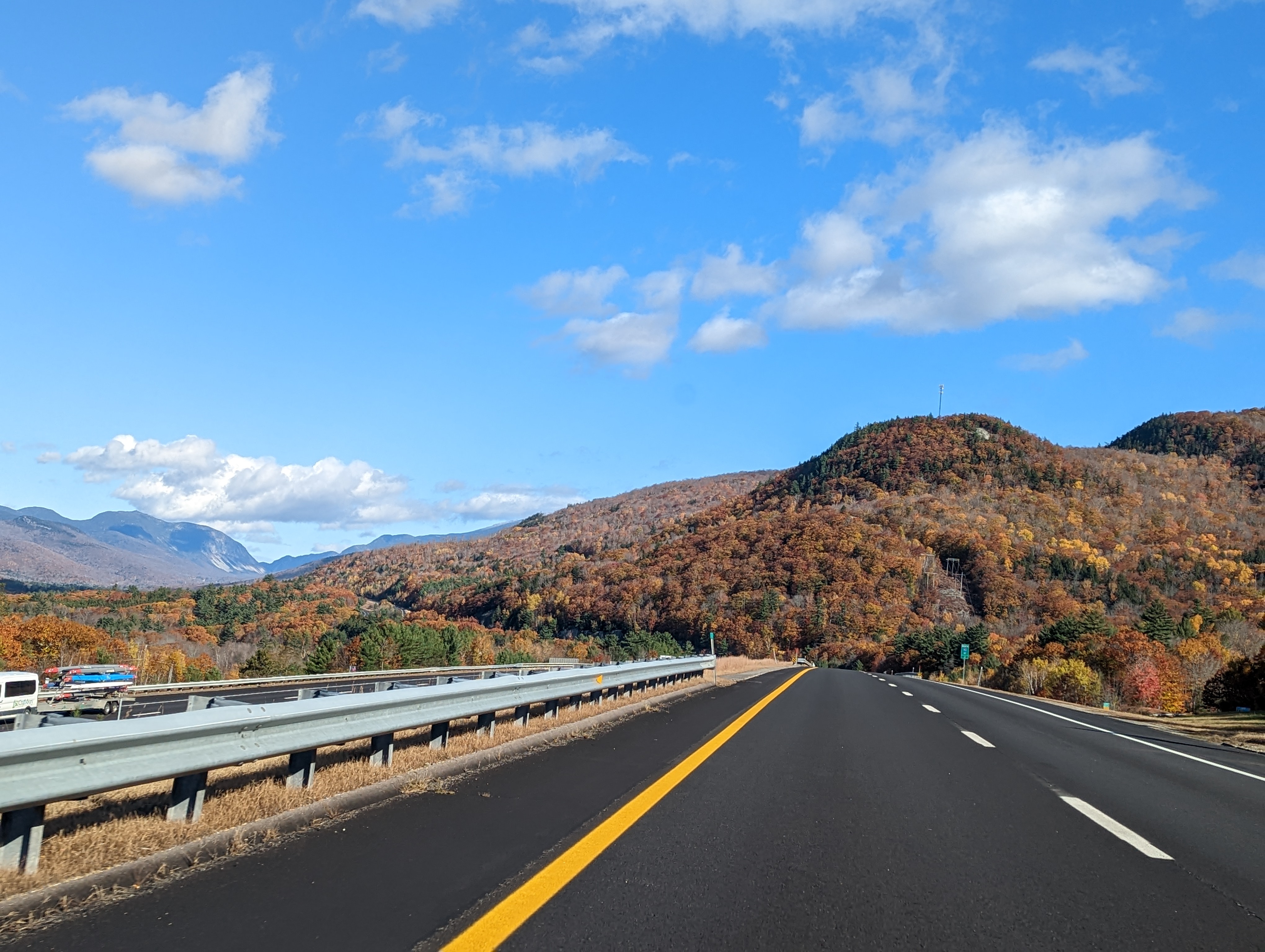 A scenic drive on a clear autumn day through the White Mountains, New Hampshire, with a road leading towards vibrant fall foliage and a mountainous horizon under a blue sky with fluffy clouds.