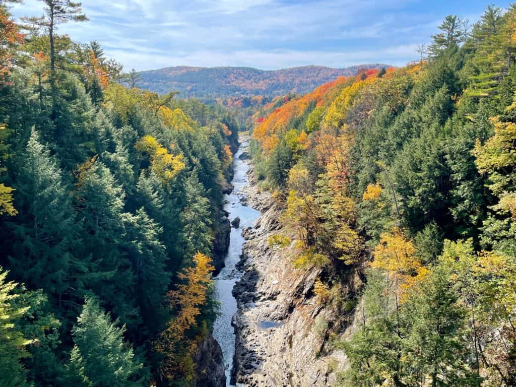 The winding Ottauquechee River cuts through a colorful autumn landscape in Woodstock, Vermont, with a mix of evergreen and deciduous trees in mid-fall splendor.