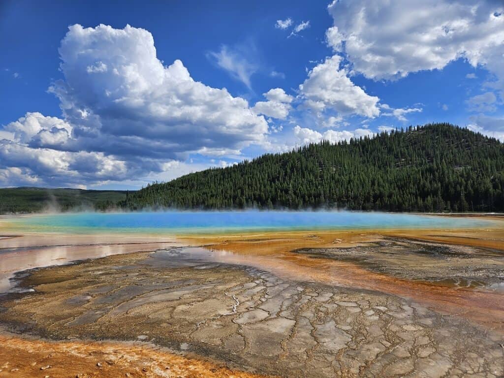 The Grand Prismatic Spring at Yellowstone National Park, Wyoming, with its striking, vividly colored geothermal pool transitioning from turquoise blue to orange, contrasted against the rustic geothermal patterns and a lush forest backdrop under a dynamic sky.