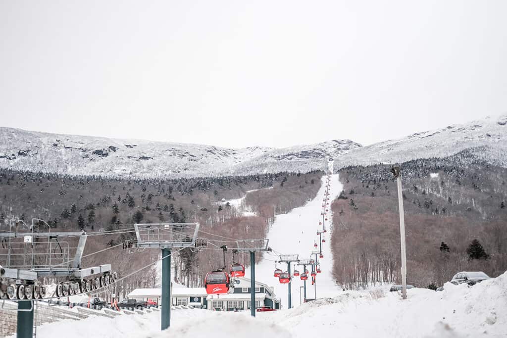 A serene winter landscape at Stowe, Vermont, featuring a ski lift with red cabins ascending towards a snow-covered mountain peak, with dense snow-dusted forests on either side.