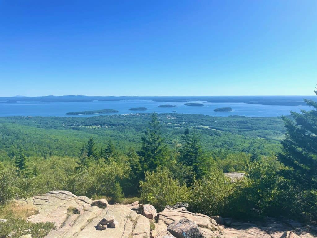 Scenic overlook from a mountaintop in Bar Harbor, Maine, showcasing a panoramic view of the Atlantic Ocean, lush greenery, and distant islands on a clear day with vibrant blue skies.