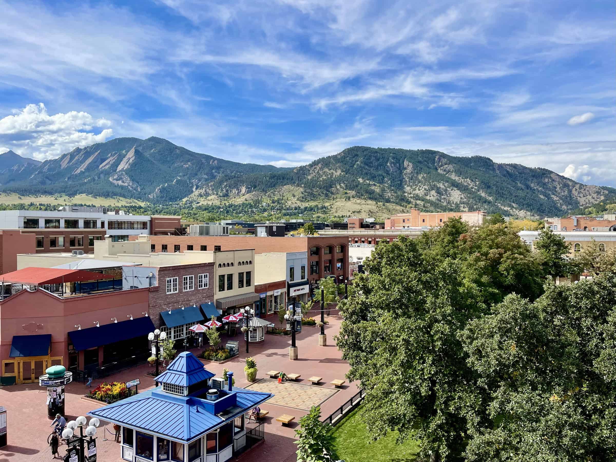 A picturesque view of Boulder, Colorado, with a vibrant cityscape in the foreground featuring distinctive architecture and a public square, set against the stunning backdrop of the Rocky Mountains under a clear blue sky.
