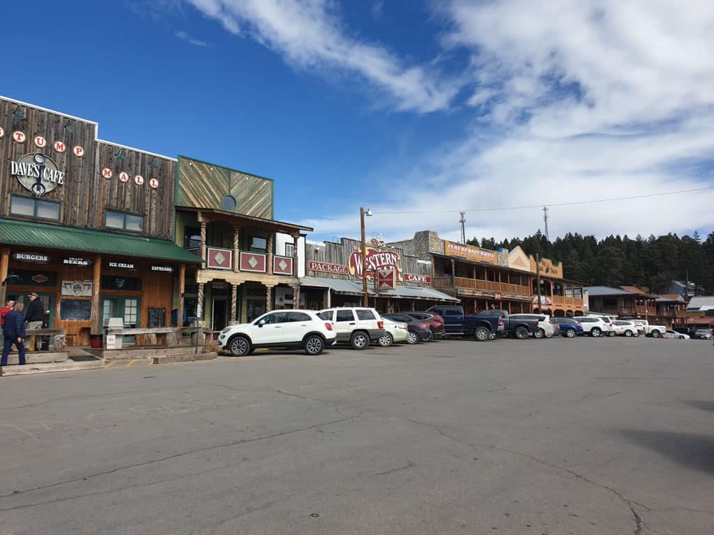 A bustling street scene in Cloudcroft, New Mexico, showcasing rustic storefronts with wooden facades, including 'Dave's Café' and a 'Western Bar,' under a clear blue sky, with parked cars lining the street