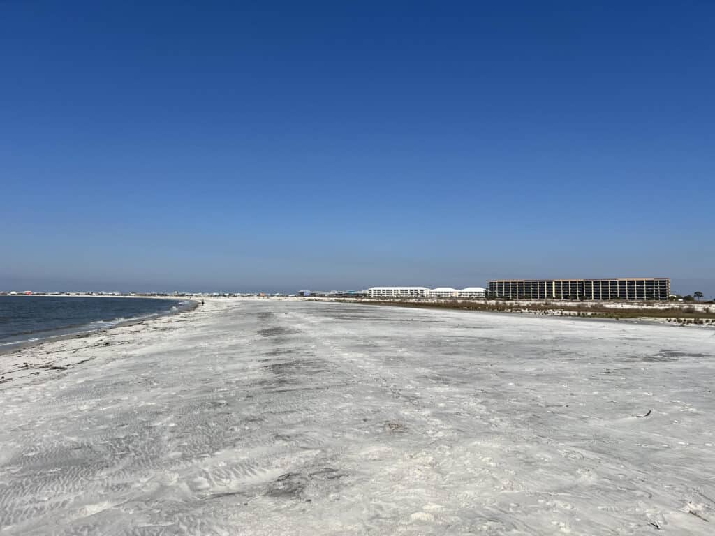 A serene beachscape of Dauphin Island, Alabama, with its unique white sandy shoreline leading to distant waterfront properties under a clear blue sky.