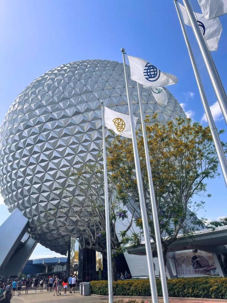 Iconic view of Spaceship Earth at EPCOT in Disney World, the geodesic sphere is a symbol of innovation and imagination, with fluttering flags bearing EPCOT logos in the foreground against a brilliant blue sky.