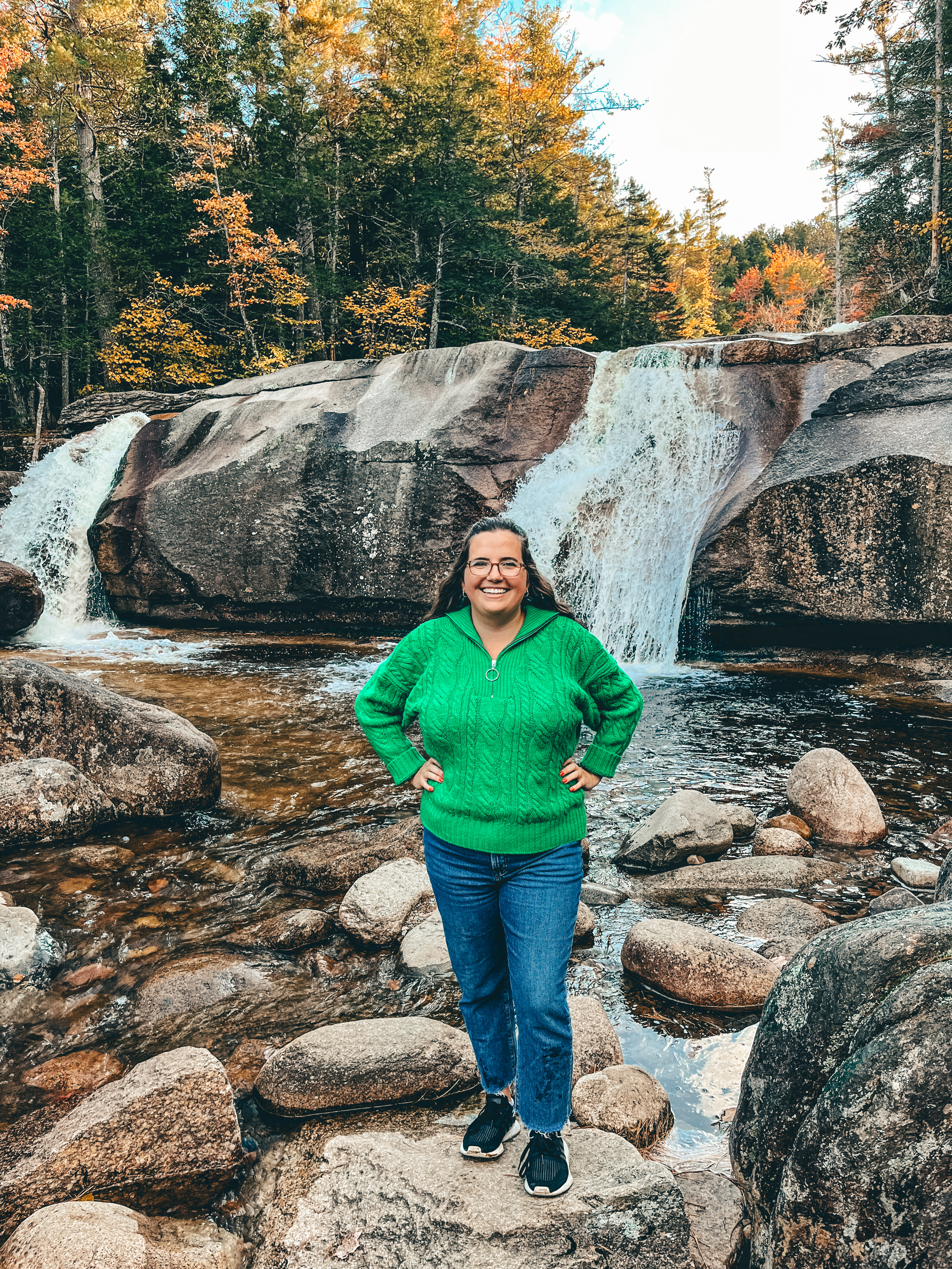 A woman in a bright green sweater and blue jeans stands smiling in front of a small waterfall surrounded by autumn-colored trees, an ideal setting for showcasing a fall travel capsule wardrobe