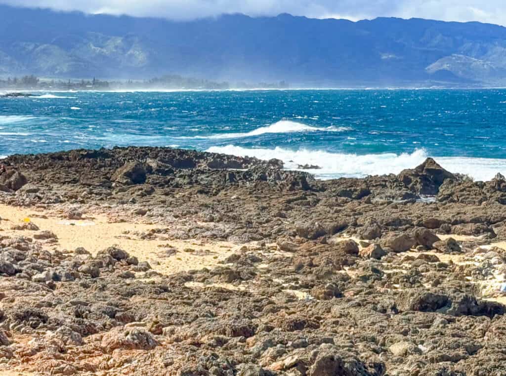 A dynamic coastal view of Oahu, Hawaii, with rugged, rocky terrain in the foreground leading to vibrant blue ocean waves, against a backdrop of distant mountains partially obscured by mist