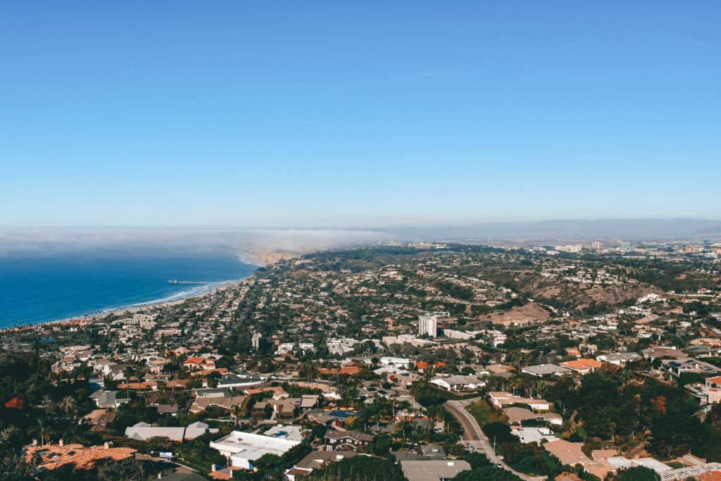An expansive aerial view of San Diego, California, showing a dense layout of residential and commercial buildings leading towards a shoreline dotted with numerous boats, all under a clear blue sky with a distant fog over the ocean.