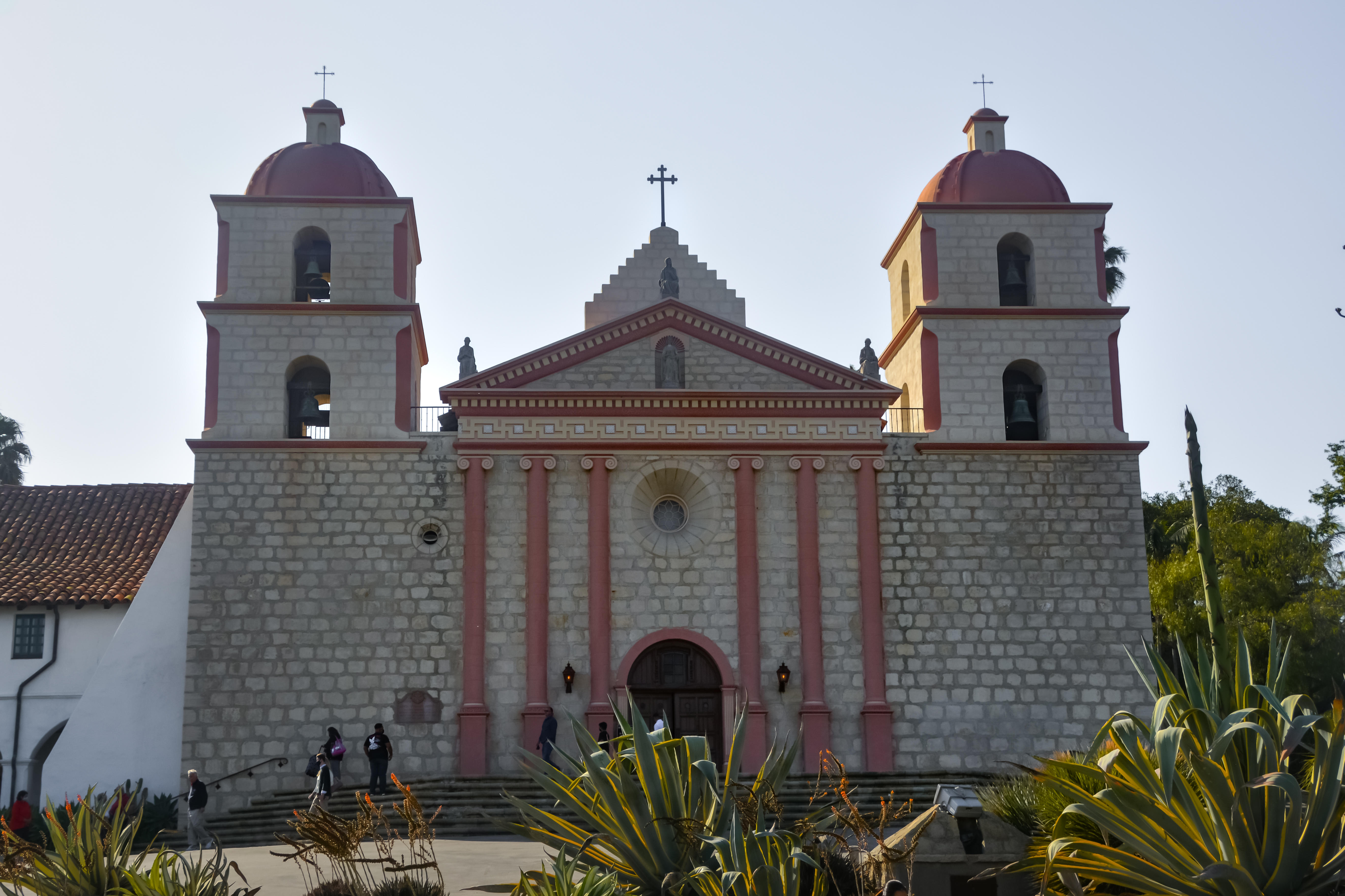 The historic Mission Santa Barbara in California, showcasing its distinctive twin bell towers with red-tiled domes, stone facade, and grand entrance flanked by statues, captured on a sunny day with vibrant greenery in the foreground