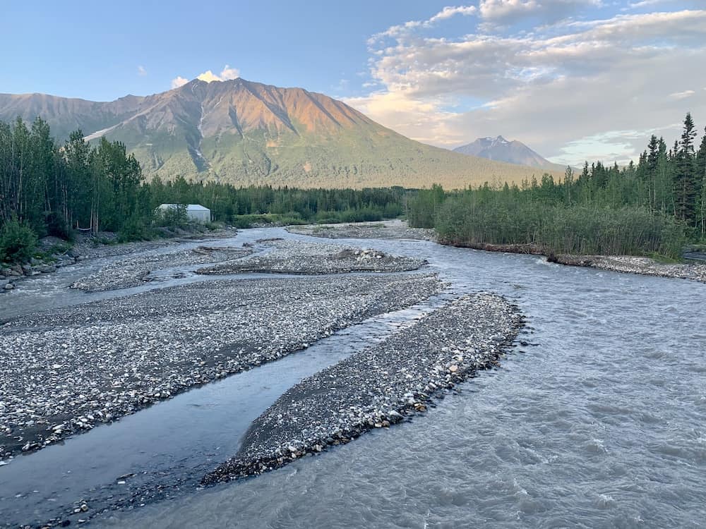 The serene landscape of Wrangell-St. Elias National Park in Alaska, featuring a meandering river with rocky banks, lush greenery, and majestic mountains with peaks touched by the light of the setting sun