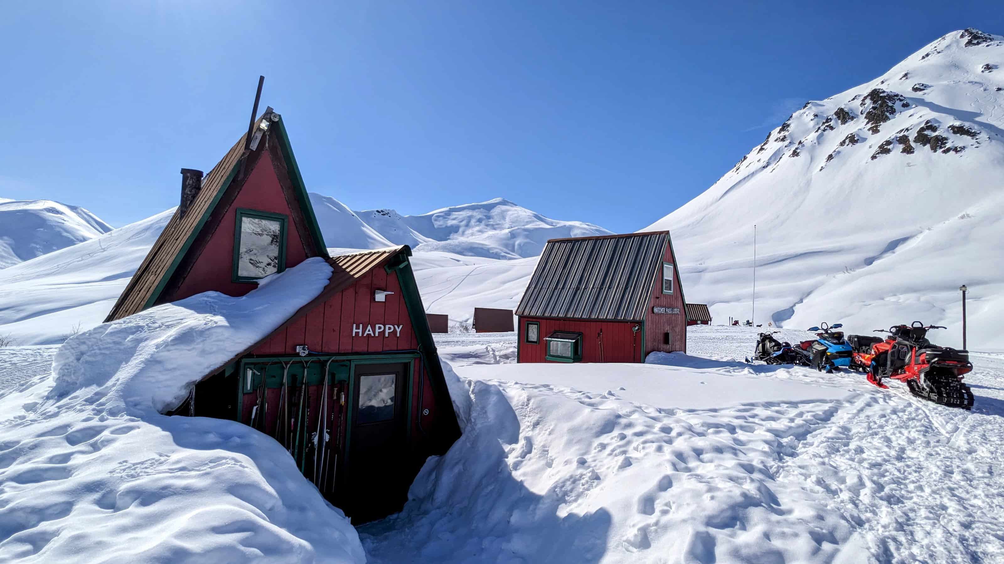 A vibrant red, A-frame cabin with the word 'HAPPY' above the door, nestled in a snowy landscape with snow-covered mountains in the background and a row of parked snowmobiles to the side under a clear blue sky