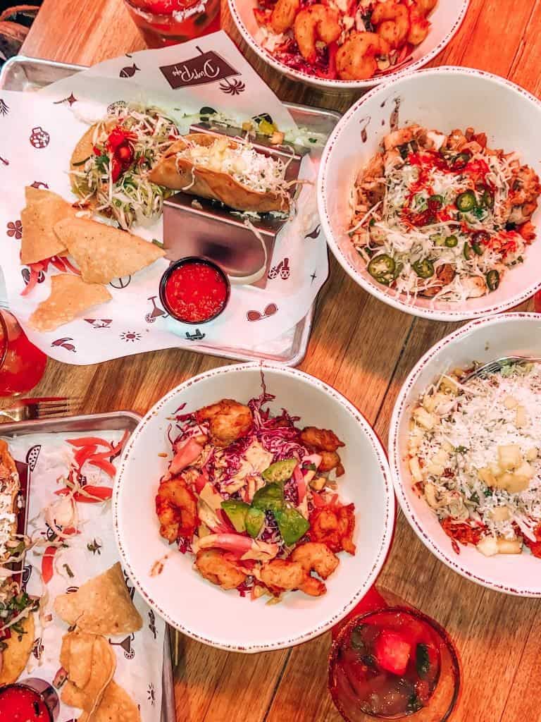 A vibrant array of Mexican dishes from Diego Pops, featuring colorful bowls of spicy shrimp, fresh salads, and taquitos, alongside crispy tortilla chips with red salsa, all presented on a wooden table for a mouth-watering meal.