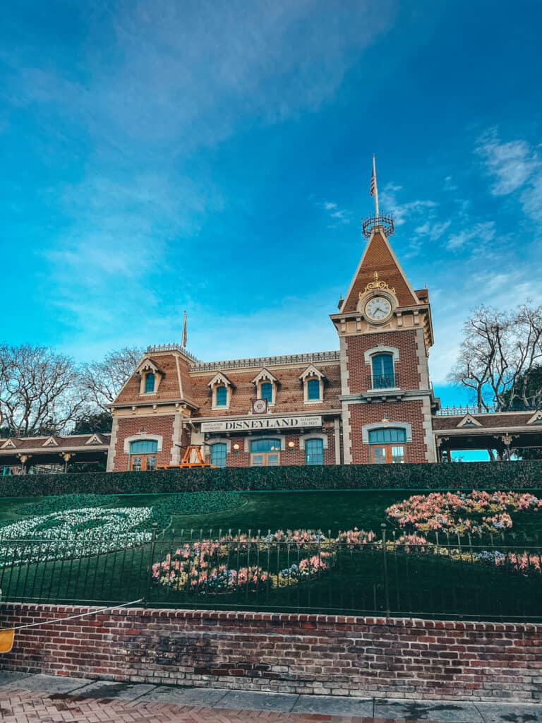 The classic Disneyland Railroad station, with its charming brick facade and clock tower, stands under a clear blue sky. A well-manicured floral display in the shape of Mickey Mouse's head welcomes visitors at the entrance.
