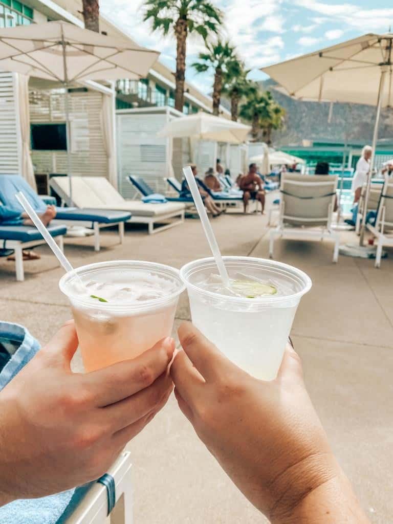 Close-up of two hands holding refreshing cocktails by the poolside at Mountain Shadows resort, with a backdrop of loungers, umbrellas, and palm trees against a mountainous landscape, capturing a leisurely moment of relaxation.